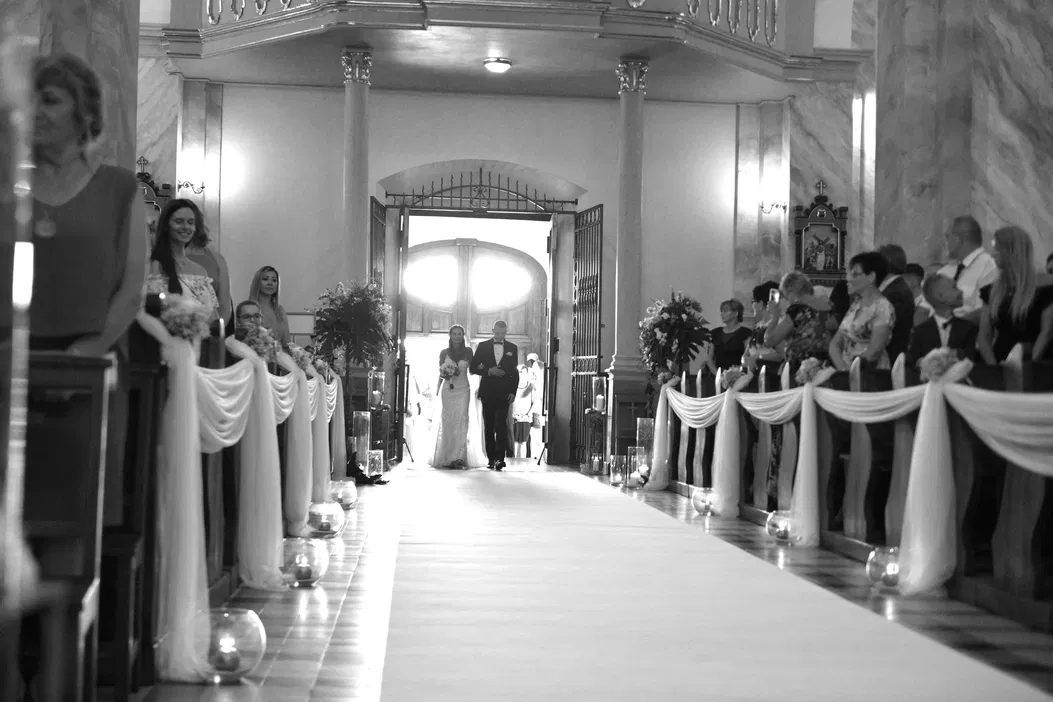 Czarno-białe zdjęcie ceremonii ślubnej w kościele, panna młoda w sukni, goście w ławkach, ozdobne dekoracje i świece na podłodze.