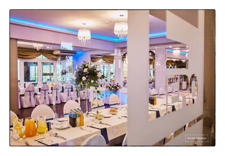  Elegancka sala weselna z dekoracjami, stoły ustawione na przyjęcie, krzesła pokryte białymi pokrowcami z niebieskimi kokardami, w tle bukiety kwiatów i żyrandole. 