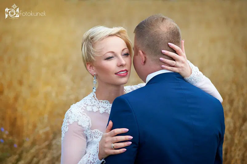  Para młoda w romantycznym uścisku na tle pola zboża, panna młoda w eleganckiej koronkowej sukni ślubnej i pan młody w granatowym garniturze, naturalna sesja zdjęciowa w plenerze. 