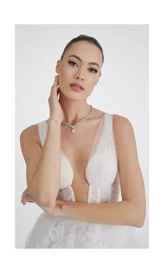  Modelka w eleganckiej, białej sukni wieczorowej z transparentnymi elementami, prezentująca elegancką biżuterię w postaci naszyjnika i bransoletki, na neutralnym tle. 