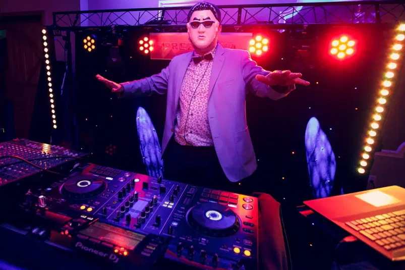  DJ w masce i okularach przeciwsłonecznych przy konsoli DJ-skiej na kolorowej imprezie z efektami świetlnymi. 