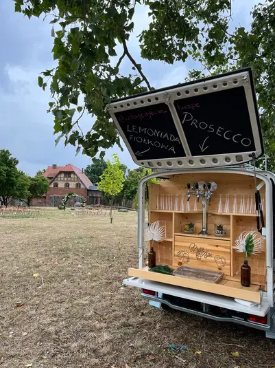  Mobilny bar z prosecco i lemoniadą fiołkową na tle rustykalnego domu i ogrodu, idealny na letnie przyjęcia na świeżym powietrzu. 