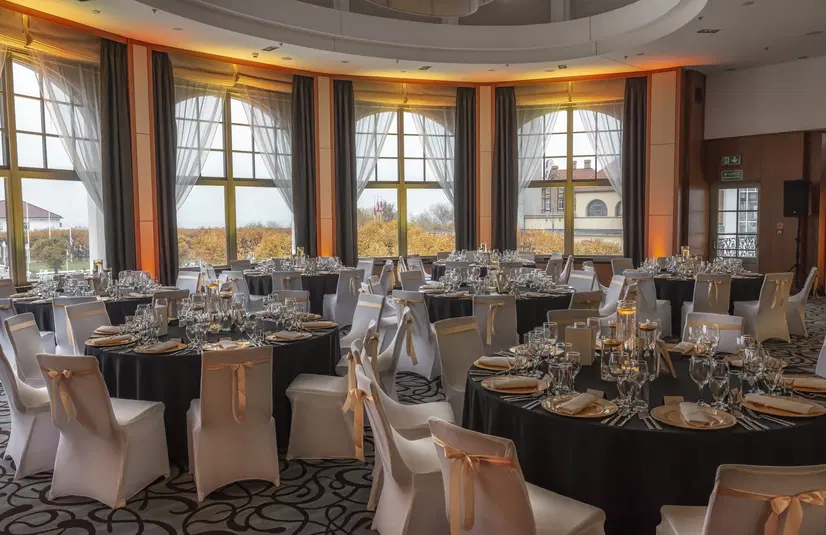  Elegancka sala bankietowa z zapleczem cateringowym, udekorowana na przyjęcie weselne, okrągłe stoły z białymi pokrowcami na krzesłach i złotymi akcentami, oświetlenie w oknach z widokiem na ogród. 