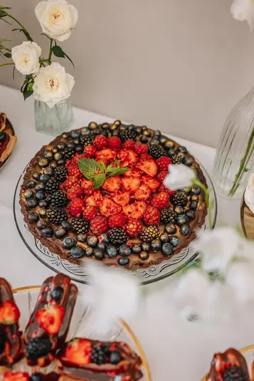  Tort czekoladowy udekorowany owocami leśnymi, takimi jak maliny, jagody i jeżyny, na eleganckim talerzu, z białymi różami w tle. 