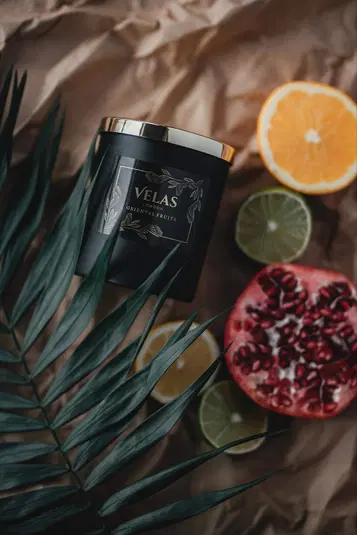  Czarna świeca zapachowa Velas z napisem Oriental Fruits na złotym tle, otoczona przez egzotyczne owoce jak pomarańcza, limonka i granat, na tle zielonych liści i papieru do pakowania. 
