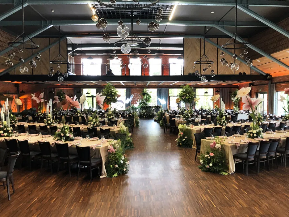 Sala weselna z dekoracjami kwiatowymi, elegancko nakrytymi stołami, otwartą przestrzenią i industrialnym wykończeniem.