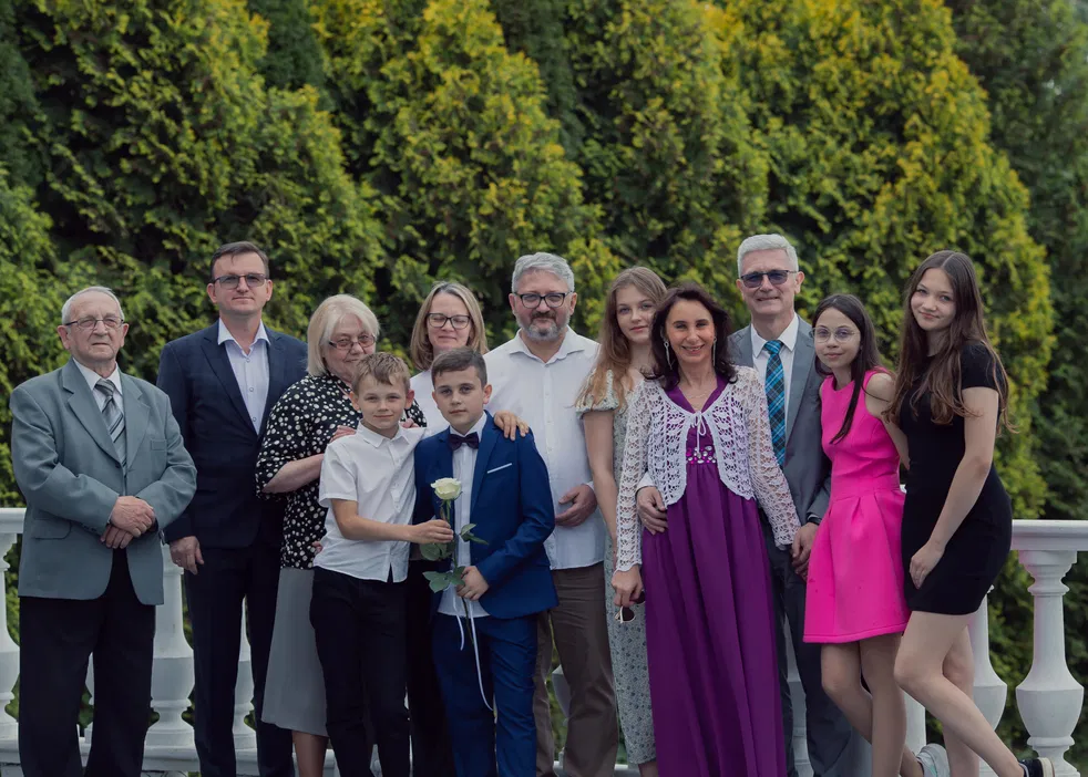 Grupa osób pozujących do zdjęcia na tle zielonego żywopłotu, elegancko ubrani, uśmiechnięci, w różnych stylizacjach formalnych i casualowych, w ciepły dzień na zewnątrz.