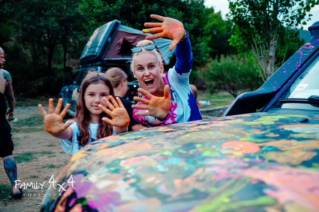 Dwie osoby z pomalowanymi dłońmi stoją przed kolorowo pomalowanym samochodem terenowym na tle lasu, Family 4x4 Adventure.