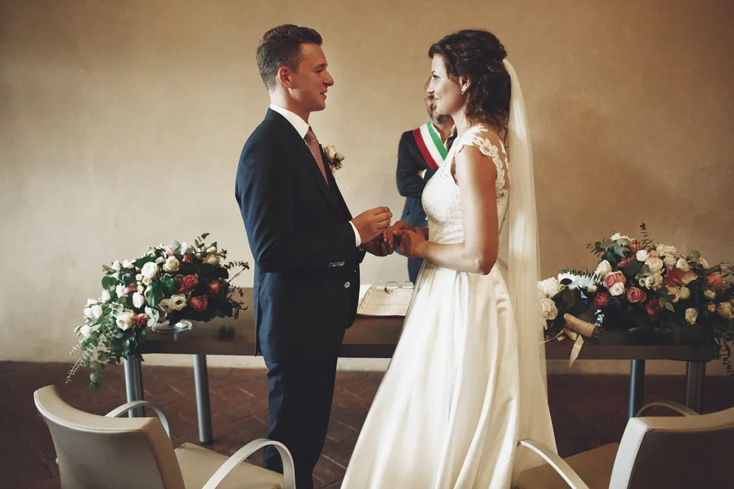 Para młoda podczas ceremonii ślubnej wymienia obrączki w eleganckiej sali, ozdobionej bukietami kwiatów, klasyczna suknia ślubna i garnitur, romantyczna atmosfera.