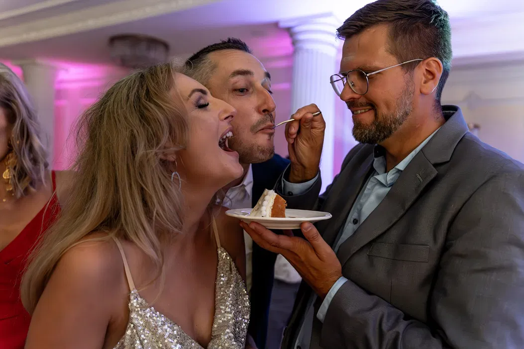 Trójka ludzi na weselu, zabawa z tortem weselnym, eleganckie stroje, przyjęcie w jasnym oświetleniu, kolumny w tle, radosna atmosfera.
