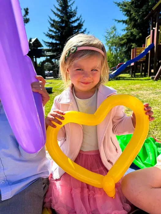 Dziewczynka z blond włosami trzymająca żółty balon w kształcie serca na tle placu zabaw, słoneczny dzień.