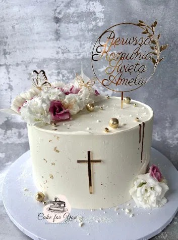 Tort na Pierwszą Komunię Świętą z kremowymi kwiatami, złotym krzyżem i personalizowanym topperem.