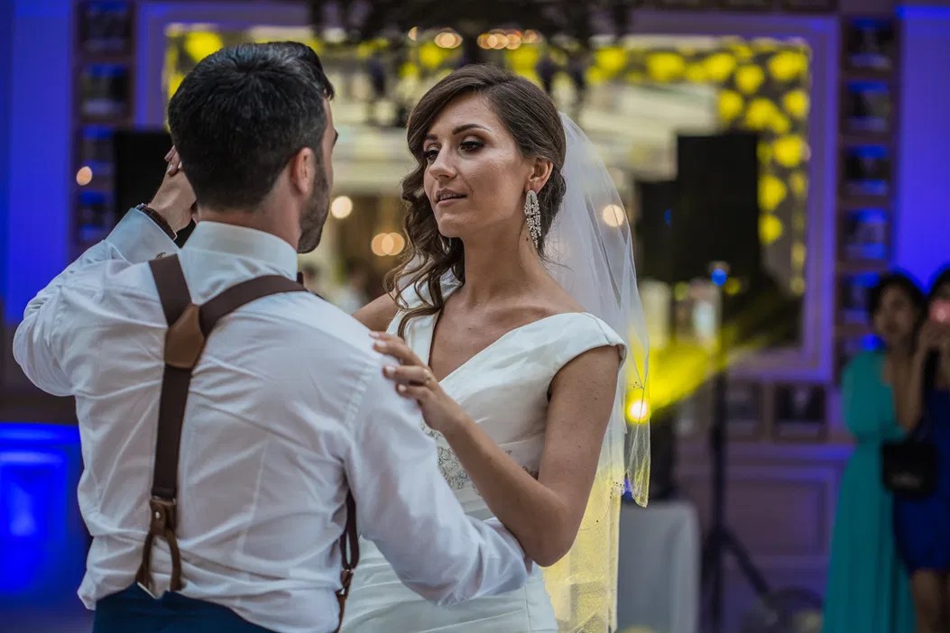 Para młoda tańcząca pierwszy taniec na eleganckim weselu, panna młoda w białej sukni z welonem, pan młody w białej koszuli z szelkami.