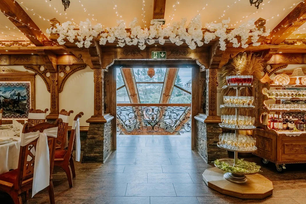 Wnętrze stylowej restauracji z drewnianymi dekoracjami, ozdobnymi krzesłami i stołem, elegancko nakrytymi na przyjęcie, z widokiem na ozdobny balkon i zielony krajobraz.