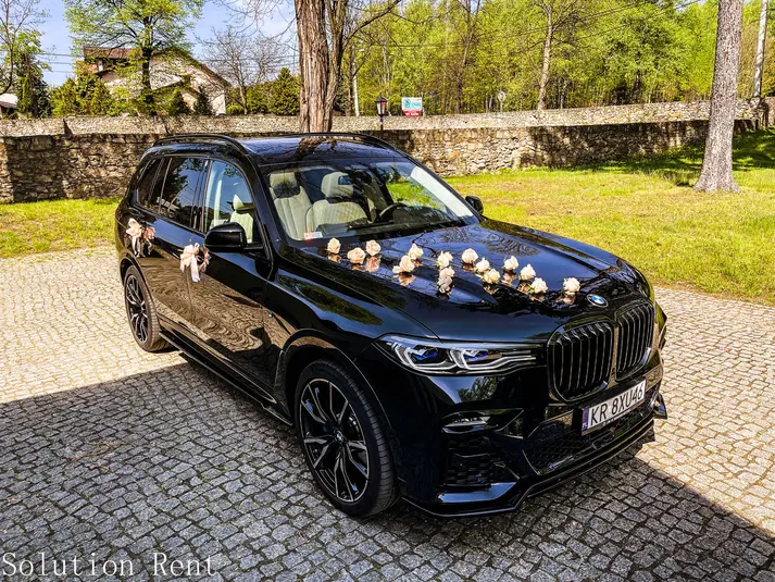  Luksusowy czarny samochód BMW SUV ozdobiony kwiatami na ślub, stojący na brukowanej powierzchni w zielonym otoczeniu. Idealny wybór na wynajem auta do ślubu. 