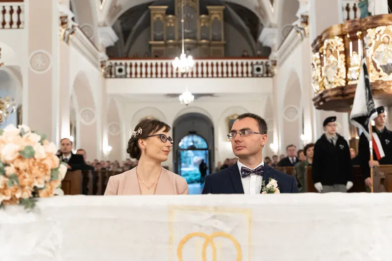 Para młoda podczas ceremonii ślubnej w eleganckim kościele, z dekoracjami kwiatowymi i uczestnikami w tle. 