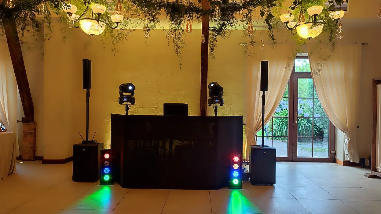 Stanowisko DJ-a w eleganckiej sali weselnej z kolorowymi światłami i ozdobami roślinnymi.