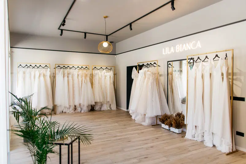  Wnętrze eleganckiego salonu ślubnego z sukniami ślubnymi na wieszakach, nowoczesne oświetlenie, drewniana podłoga, zielone rośliny jako dekoracja. 