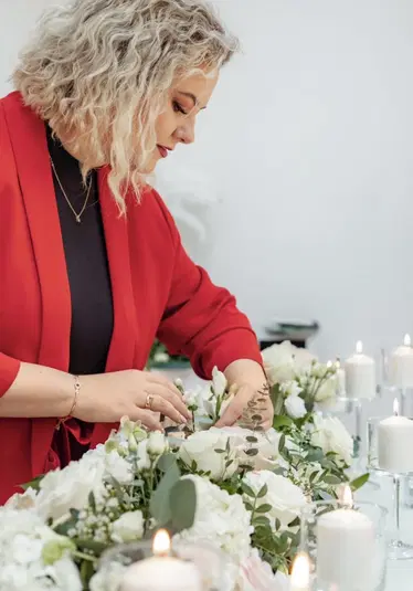  Kobieta w czerwonym płaszczu tworzy kompozycję kwiatową z białych kwiatów i zieleni, otoczona przez zapalone świece, wnętrze jasne i eleganckie. 