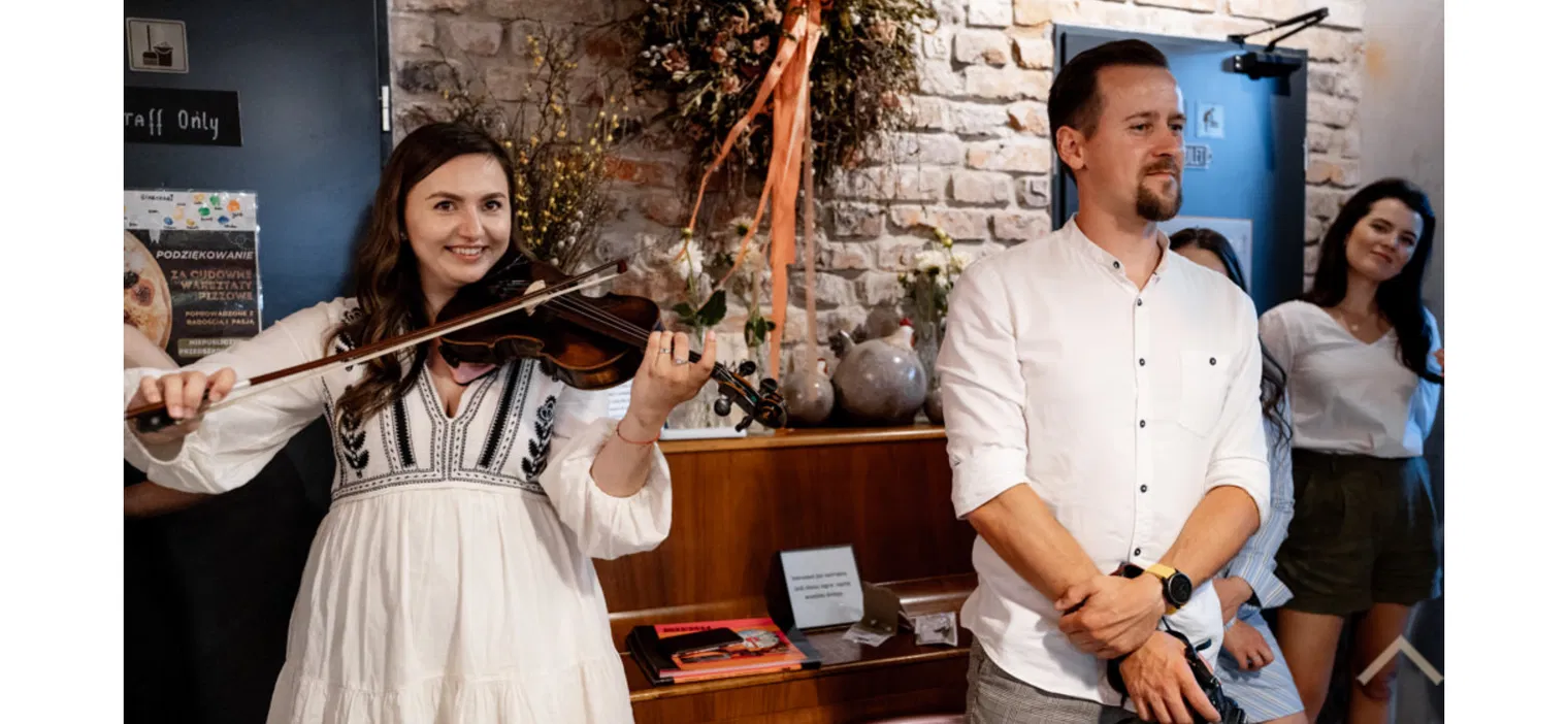 Muzyk grający na skrzypcach podczas uroczystości wewnętrznej z grupą ludzi w tle, elegancka atmosfera, dekoracje kwiatowe na ścianie.
