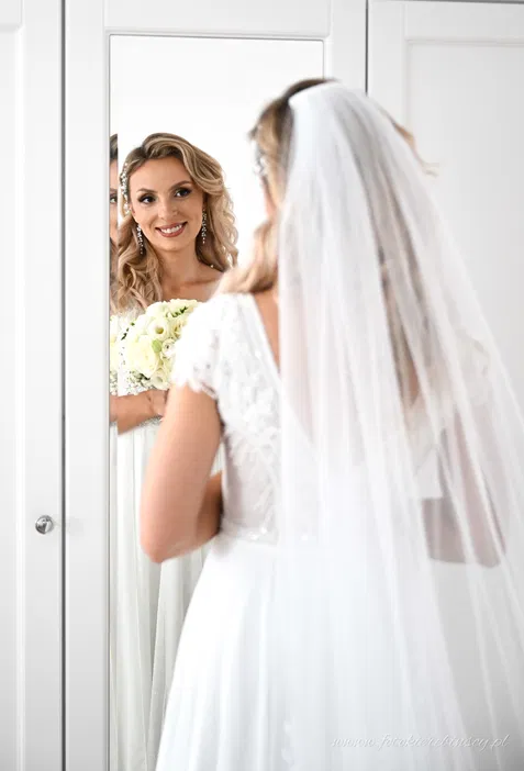 Panna młoda w sukni ślubnej przed lustrem, ubrana w welon, trzyma bukiet białych róż, elegancka fryzura i biżuteria ślubna.