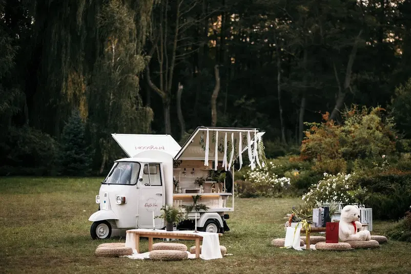  Mini food truck w lesie, ozdobiony białymi wstążkami i zielonymi liśćmi, z przytulnym miejscem do siedzenia i dużym misiem, idealne na przyjęcie plenerowe. 