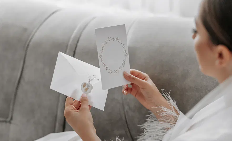  Osoba trzymająca w dłoni elegancką kartkę z wzorem wieńca oraz kopertę, siedząca na sofie w stylu glamour. 