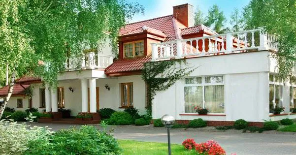 Duży, elegancki dom jednorodzinny z czerwonym dachem, otoczony zielenią i ogrodem kwiatowym, na tle błękitnego nieba.