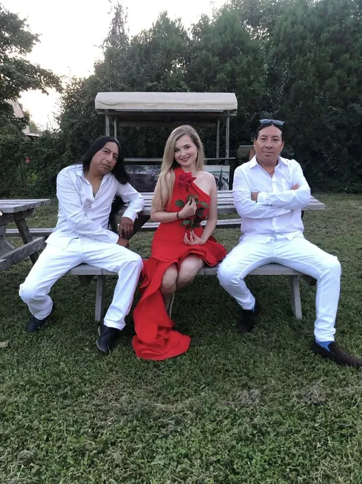 Three people sitting on a wooden bench outside, two men in white shirts and pants flanking a woman in a red dress holding a rose, with greenery in the background.
