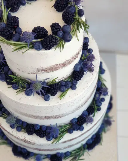  Tort weselny typu naked cake ozdobiony świeżymi jagodami, jeżynami i zielonymi gałązkami rozmarynu. 