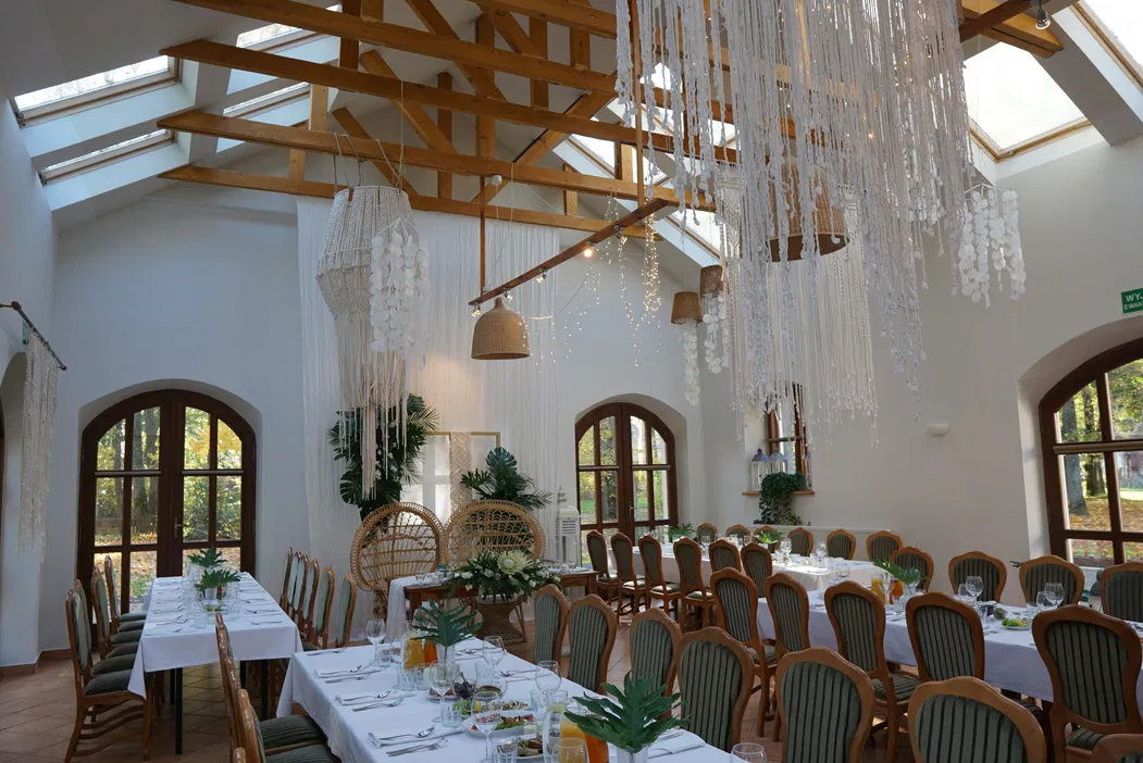 Dekorowana sala weselna z drewnianym sufitem, stołami ustawionymi wzdłuż okien, ozdobami z makramy i roślinami, eleganckie aranżacje stołów.
