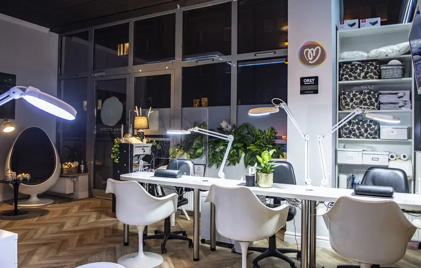 Stylowy salon kosmetyczny z nowoczesnym wyposażeniem, eleganckimi białymi fotelami i lampami do manicure, roślinami dekoracyjnymi oraz przytulnym, profesjonalnym wystrojem wnętrza. 