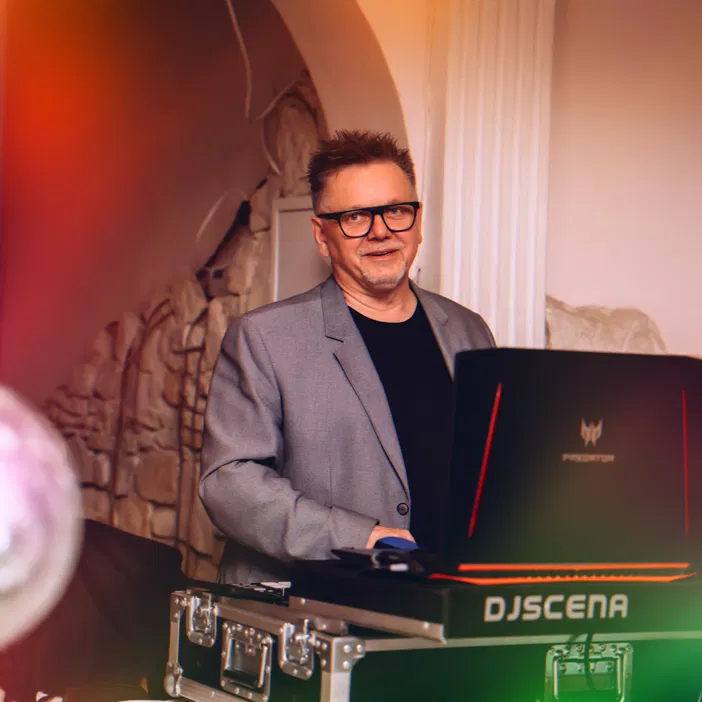 Osoba stojąca przy konsoli DJ-skim, uśmiecha się, nosi szary garnitur i okulary, w tle ozdobna ściana, efektowne oświetlenie i kolorowe refleksy świetlne.