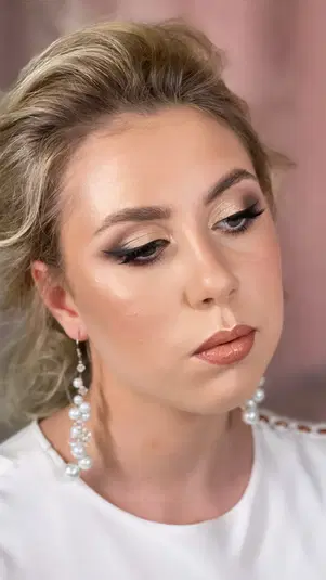  Blondynka z eleganckim makijażem wieczorowym, wyraziste oczy z czarną kreską, brzoskwiniowe usta, fryzura z upięciem i długie kolczyki z perełkami. 