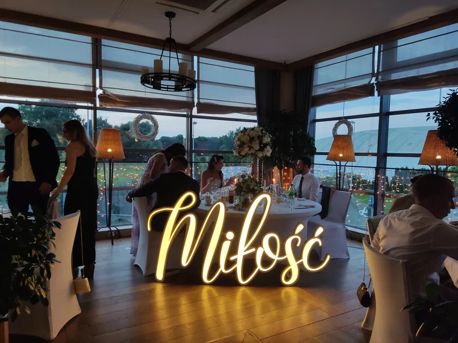 Wesele w eleganckiej sali z napisem Miłość w świetle neonowym, dekoracje kwiatowe i wieczorna atmosfera.