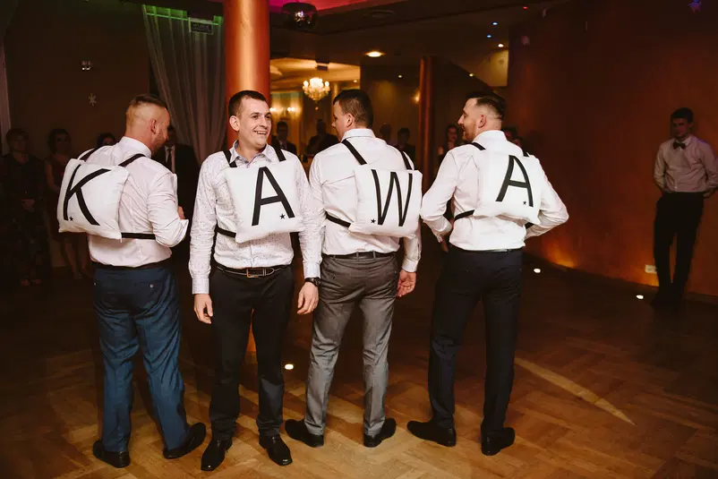  Mężczyźni na weselu z poduszkami na plecach tworzącymi napis KAWA. Impreza taneczna w eleganckim wnętrzu. 