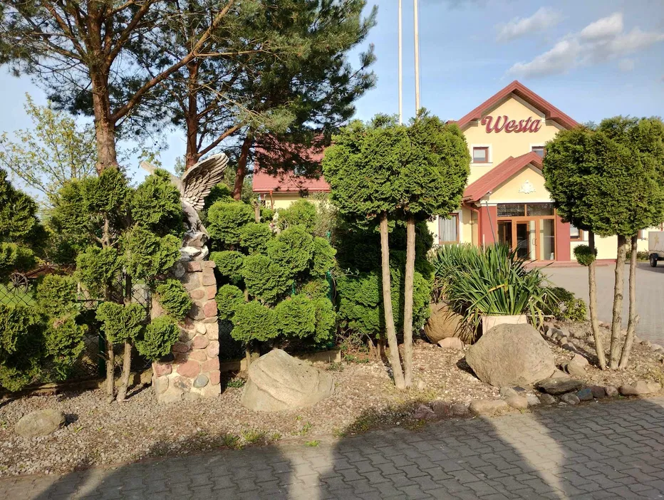 Hotel Westa z ogrodem ozdobionym rzeźbą orła, otoczony przez starannie przycięte krzewy i drzewa iglaste.