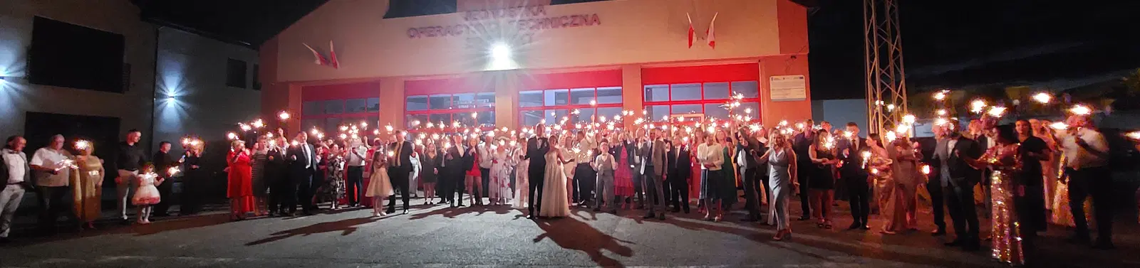  Grupa osób trzymająca zimne ognie przed budynkiem OSP, nocna scena, uroczystość, polskie flagi. 