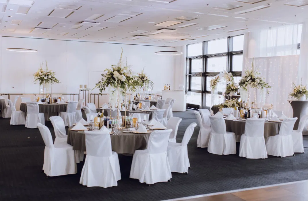 Sala bankietowa z elegancko udekorowanymi stołami, białe pokrowce na krzesła, centralne kompozycje kwiatowe, jasne wnętrze z dużymi oknami.