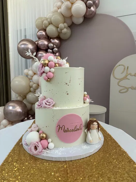 Dwupiętrowy tort na chrzest zdobiony różowymi i złotymi akcentami, otoczony balonami w pastelowych kolorach na złotym obrusie, z figurką dziecka na przyjęcie.