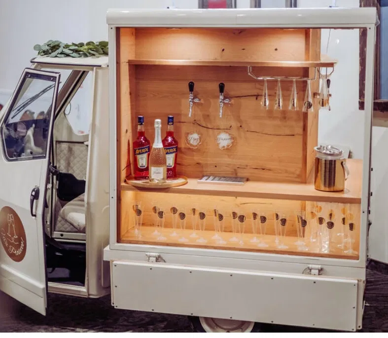 Mobilny drink bar na kółkach z drewnianym wnętrzem, butelkami Aperol i kieliszkami, idealny na eventy i wesela.