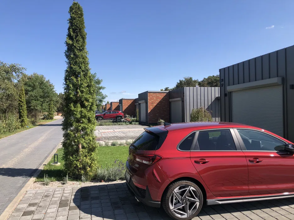 Czerwony samochód kompaktowy zaparkowany przed nowoczesnym domem na skraju ulicy, otoczony zielenią i drzewami, w słoneczny dzień.